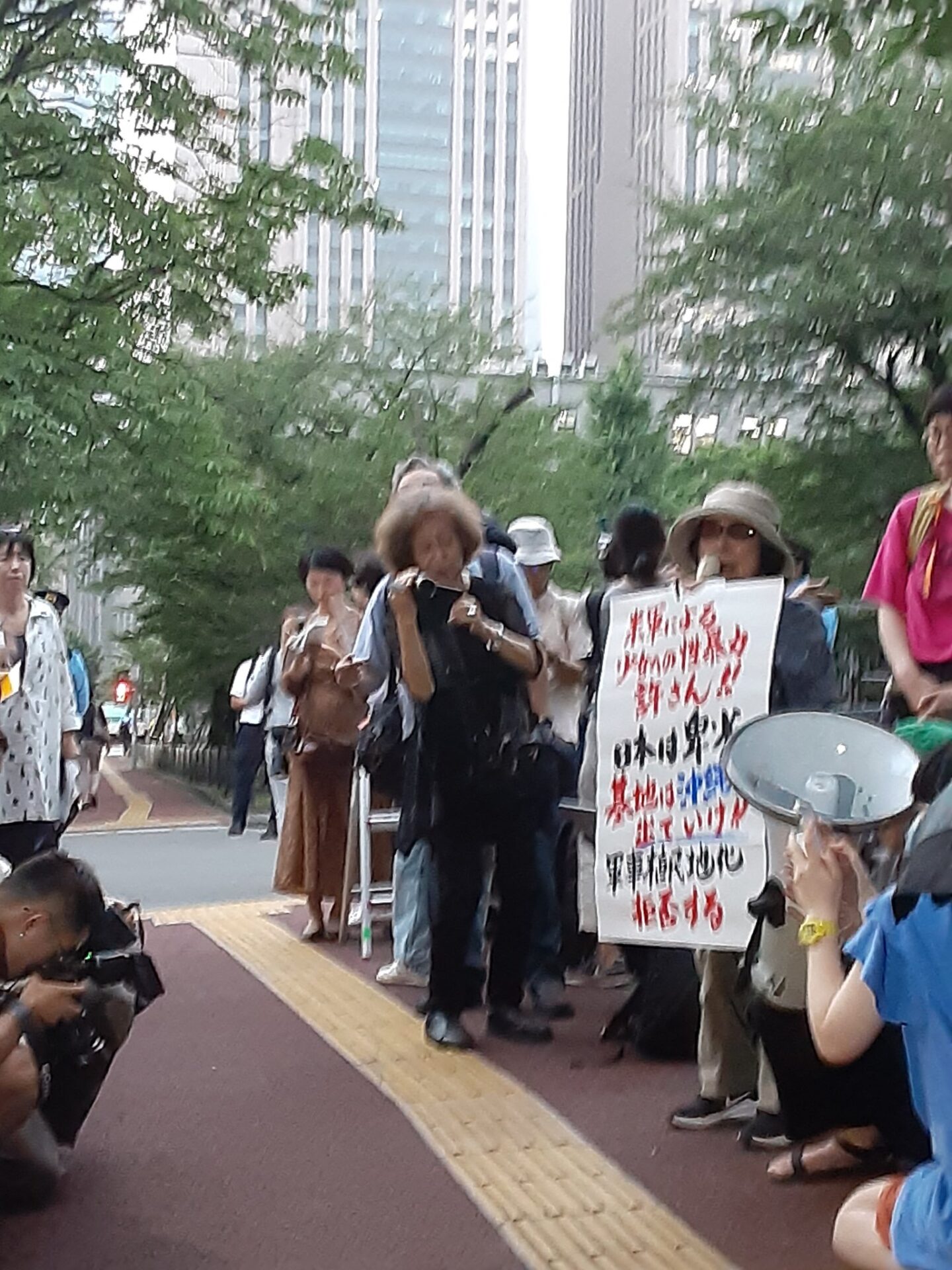 青木初子さんのスピーチの様子。「米軍による少女への性暴力許さん！日本は卑劣／基地は沖縄から出て行け！！／軍事植民地化拒否する」と書かれた大きなポスターを掲げながらマイクを握る。